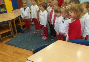 Dzieci z nauczycielką w postawie na baczność śpiewają Hymn Narodowy.